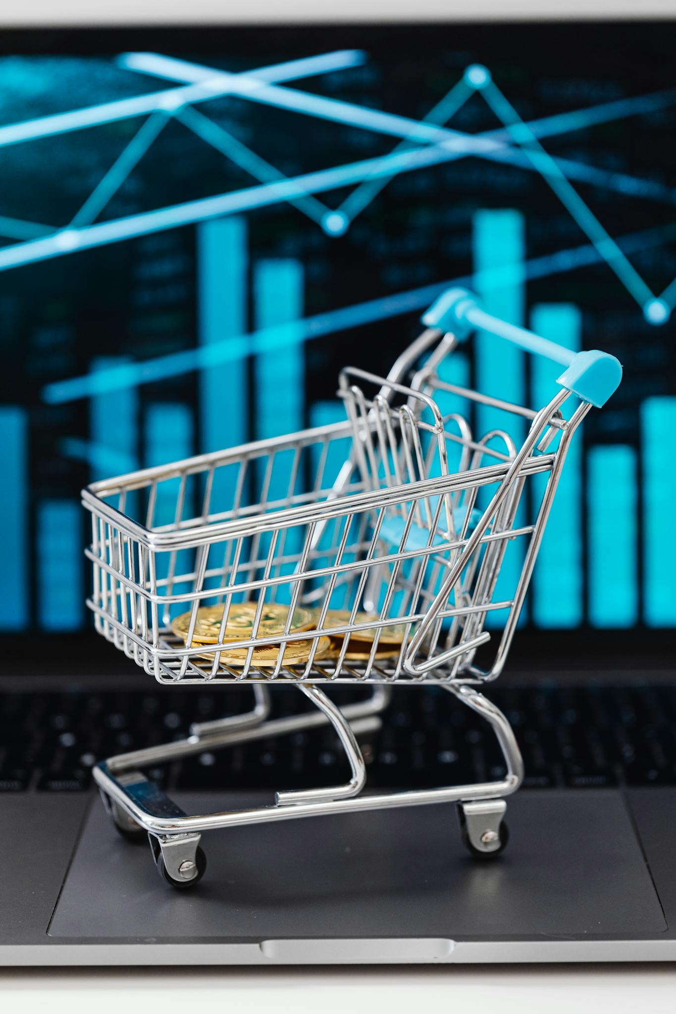 Miniature shopping cart on laptop keyboard with bitcoins, representing online trading and economy.